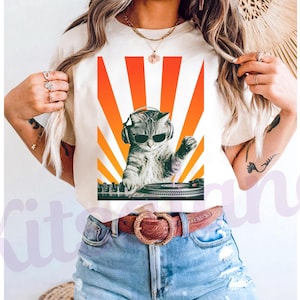 May include: Cream-colored t-shirt featuring a graphic of a cat wearing sunglasses and headphones, DJing with a turntable. The design includes a vibrant orange and yellow sunburst background. The cat is gray and white.