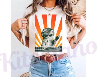 May include: Cream-colored t-shirt featuring a graphic of a cat wearing sunglasses and headphones, DJing with a turntable. The design includes a vibrant orange and yellow sunburst background. The cat is gray and white.