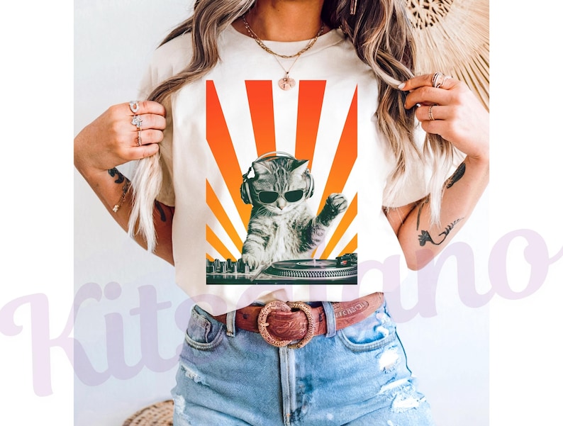 May include: Cream-colored t-shirt featuring a graphic of a cat wearing sunglasses and headphones, DJing with a turntable. The design includes a vibrant orange and yellow sunburst background. The cat is gray and white.