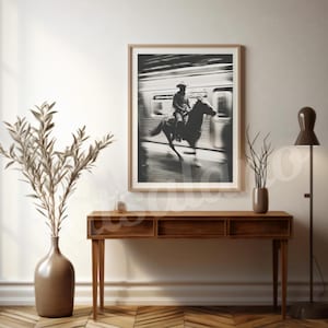May include: A framed black and white photograph depicts a cowboy on horseback, riding past a train. The image is displayed above a wooden console table, with a vase of dried plants and a floor lamp. The room has a neutral color palette.