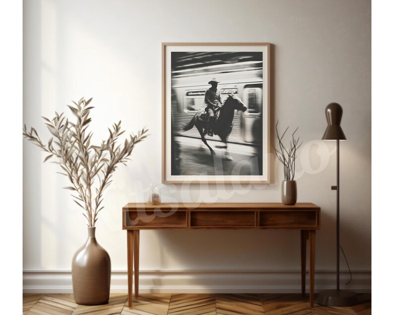 May include: A framed black and white photograph depicts a cowboy on horseback, riding past a train. The image is displayed above a wooden console table, with a vase of dried plants and a floor lamp. The room has a neutral color palette.