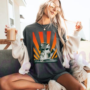 May include: Dark gray t-shirt featuring a cat DJ wearing sunglasses and headphones, set against a vibrant orange and yellow sunburst design. The cat is positioned behind a turntable. The shirt is a casual wear item.