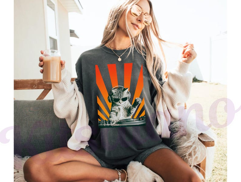 May include: Dark gray t-shirt featuring a cat DJ wearing sunglasses and headphones, set against a vibrant orange and yellow sunburst design. The cat is positioned behind a turntable. The shirt is a casual wear item.