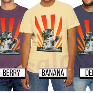 May include: T-shirts in berry, banana, and denim colors, each featuring a graphic of a cat wearing sunglasses and headphones, DJing with a turntable against a red and orange sunburst design. The shirts are displayed on models.