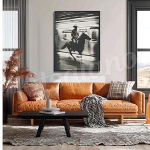 May include: Black and white framed print of a cowboy riding a horse inside a subway car. The artwork is displayed above a brown leather sofa with a black coffee table and a patterned rug.