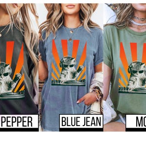 May include: T-shirts in pepper, blue jean, and moss green feature a graphic of a cat DJ wearing sunglasses and headphones, with a retro orange and yellow sunburst design. The shirts have the color names printed below.