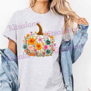 May include: A light gray t-shirt featuring a floral pumpkin design. The pumpkin is white with a brown stem and is filled with colorful flowers in shades of orange, pink, blue, and green. The text "Kitsalano" is visible on the shirt.