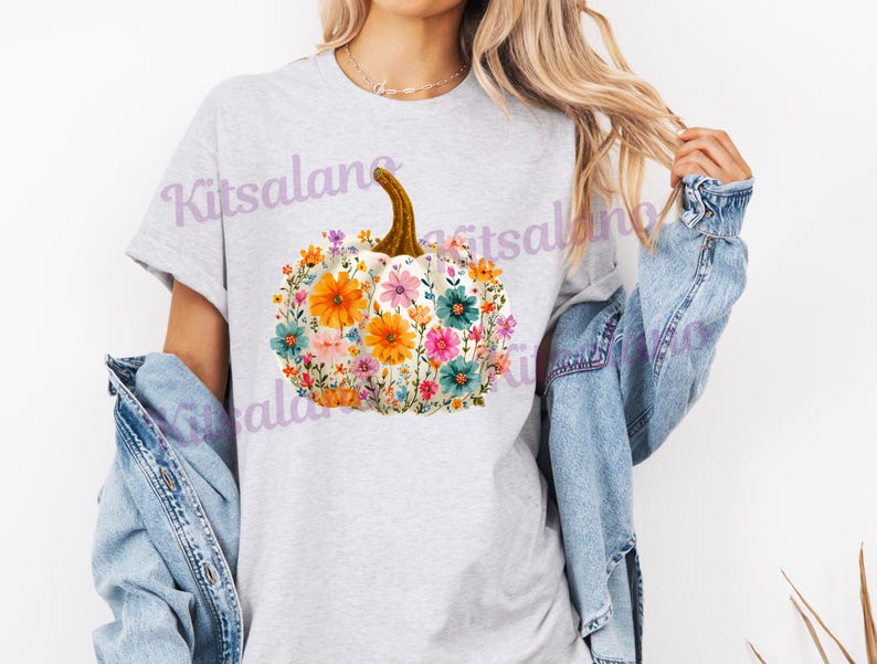May include: A light gray t-shirt featuring a floral pumpkin design. The pumpkin is white with a brown stem and is filled with colorful flowers in shades of orange, pink, blue, and green. The text "Kitsalano" is visible on the shirt.