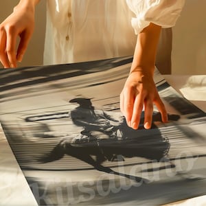 May include: Monochrome print of a cowboy riding a bucking bronco. The image is in shades of gray and sepia, with motion blur suggesting speed. The cowboy is wearing a hat and riding gear. The print is being held by a person.