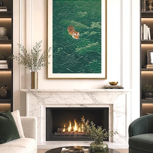 Op de afbeelding: Een ingelijste verticale wandkunstprint met een oranje en zwarte tijger die zwemt in diepgroen water met gestileerde witte golfpatronen. Het kunstwerk is ingelijst in een gouden lijst boven een open haard van wit marmer met een brandend vuur.