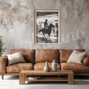 May include: A framed black and white photograph of a cowboy riding a horse, set against a blurred background. The photograph hangs above a brown leather sofa with beige pillows, a wooden coffee table, and decorative vases.