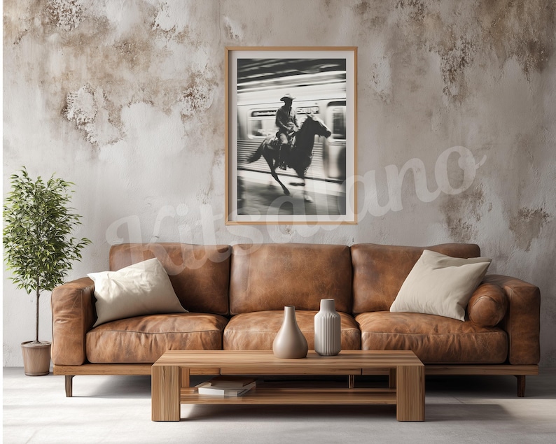 May include: A framed black and white photograph of a cowboy riding a horse, set against a blurred background. The photograph hangs above a brown leather sofa with beige pillows, a wooden coffee table, and decorative vases.