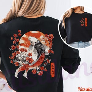 Pode incluir: Uma camisola preta de manga comprida com um gr&aacute;fico grande nas costas de um peixe koi a nadar entre flores de cerejeira vermelhas e um sol circular. Uma vers&atilde;o mais pequena do design floral com texto japon&ecirc;s est&aacute; impressa no peito.