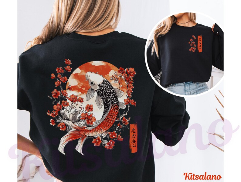 Pode incluir: Uma camisola preta de manga comprida com um gr&aacute;fico grande nas costas de um peixe koi a nadar entre flores de cerejeira vermelhas e um sol circular. Uma vers&atilde;o mais pequena do design floral com texto japon&ecirc;s est&aacute; impressa no peito.