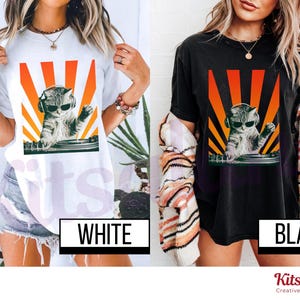 May include: White and black t-shirts featuring a graphic of a cat wearing sunglasses and headphones, DJing with a turntable. The cat graphic is set against a backdrop of orange and yellow rays. The word "WHITE" and "BLACK" are displayed on the shirts.