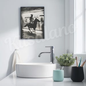 May include: A black and white framed print of a cowboy riding a horse in front of a train. The print hangs above a white sink with a chrome faucet. A small potted plant, a light blue cup, and a black cup with toothbrushes are on the counter.