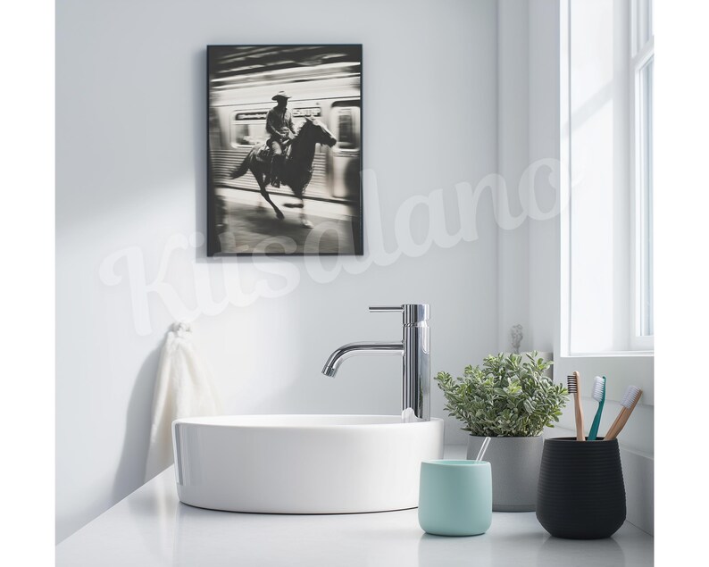 May include: A black and white framed print of a cowboy riding a horse in front of a train. The print hangs above a white sink with a chrome faucet. A small potted plant, a light blue cup, and a black cup with toothbrushes are on the counter.