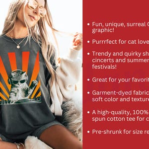 May include: Dark gray t-shirt featuring a cat DJ graphic with sunglasses and headphones, set against an orange and yellow sunburst design. The text on the shirt says "Fun, unique, surreal Cat DJ graphic!"