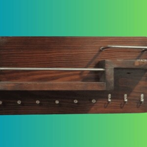 Puede incluir: Un porta llaves de madera marrón oscuro con un estante y un gancho de metal. El porta llaves tiene 8 ganchos de metal para colgar llaves.