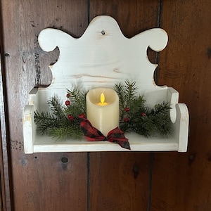 May include: A white wooden shelf with a decorative top, holding a flameless candle, evergreen branches, red berries, and a red and black plaid bow. The shelf is mounted on a dark wooden surface.