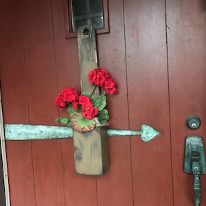 Puede incluir: Una maceta de madera con flores rojas colgando de una puerta de madera roja. La maceta está unida a la puerta con un gancho de metal.