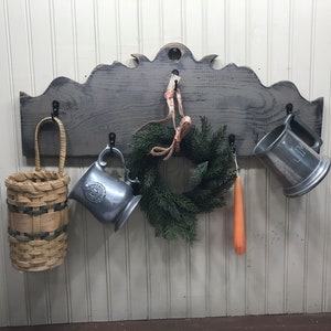 Puede incluir: Un estante de pared de madera rústico con ganchos, con un acabado de lavado gris. Tiene una parte superior decorativa con un borde festoneado. El estante está decorado con una pequeña cesta tejida, dos jarras de peltre, una pequeña corona de hoja perenne y una vela larga de color naranja.