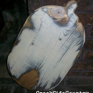 Puede incluir: Una tabla de servir de madera grande, de forma ovalada, con un acabado de pintura blanca desgastada y un asa de madera natural. La tabla tiene un estilo rústico y de granja.