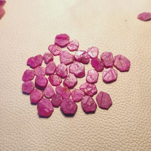 Puede incluir: Una colección de pequeñas piedras preciosas de rubí en bruto y sin pulir en varios tonos de rosa vibrante y rojo intenso. Cada piedra presenta una forma irregular y facetada con una superficie mate y texturizada, dispersas en un grupo.