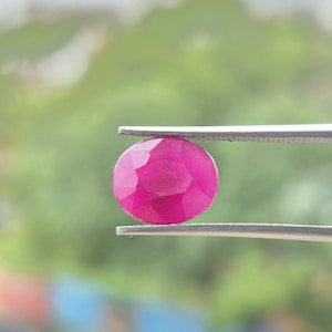 Op de afbeelding: Een levendige, ovaal geslepen robijn met een diepe roze-rode tint en meerdere gefacetteerde oppervlakken die het licht reflecteren. De edelsteen wordt stevig vastgehouden tussen de twee metalen punten van een zilverkleurige pincet.