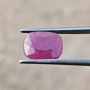 Puede incluir: Una piedra preciosa rosa de corte cojín sostenida por pinzas de metal. La piedra presenta una superficie facetada que refleja la luz, mostrando un tono magenta vibrante con claridad interna visible y un acabado pulido, asegurada entre dos brazos de metal texturizado.