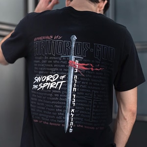 Könnte beinhalten: Schwarzes T-Shirt mit einer Grafik eines Schwertes und dem Text "Armor of God" und "Sword of the Spirit" mit einem Zitat aus Epheser 6:11. Der Text ist in weißer Schrift mit rotem und schwarzem Hintergrund.