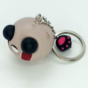 May include: A keychain featuring a round, pale pink character head with large black eyes and a red stripe. The keychain includes a silver ring and a black paw-shaped charm with pink details.