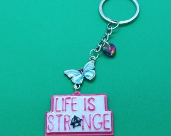 May include: A pink and white keyring with a butterfly charm and a heart charm. The keyring has the words "Life is Strange" written on it.
