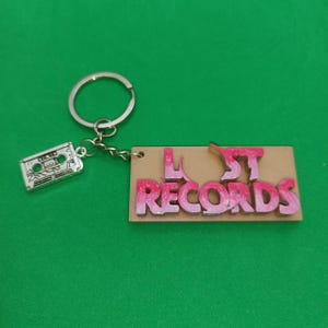 Puede incluir: Un llavero con un dije de cinta de casete plateada y una etiqueta rectangular. La etiqueta es de color canela con las palabras "LOST RECORDS" en rosa, con un llavero plateado. El fondo es verde liso.