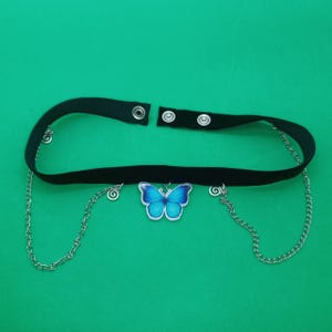May include: Black velvet choker necklace featuring a blue butterfly pendant and silver chain detailing. The choker has a snap closure and small silver charms. The butterfly pendant is light blue with darker blue detailing.