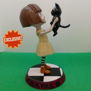 Puede incluir: Figura de una niña con un vestido amarillo sosteniendo un gato negro. La figura tiene cabello castaño, calcetines a rayas y botas naranjas. La base es a cuadros blancos y negros con el texto "FRANS &" y una silueta de gato. Una estrella naranja con la palabra "EXCLUSIVE!" está en la imagen.