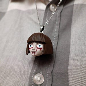 May include: A silver chain necklace with a pendant featuring a small, sculpted head. The head has brown hair, blue eyes, and red streaks under the eyes and mouth. The pendant is attached to a silver chain.
