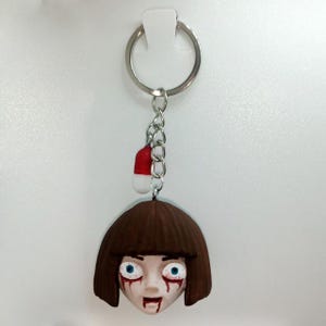 May include: A handmade keychain featuring a spooky, stylized face with brown hair, blue eyes, and red blood streaks. The keychain includes a small red and white pill-shaped charm and a silver keyring.