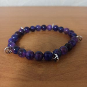 May include: A purple gemstone bracelet with silver swirl charms. The bracelet is made of round beads with a purple and white marbled pattern.
