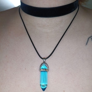 Puede incluir: Un collar gargantilla negro con un colgante de cristal azul. El cristal tiene forma de punta y tiene un engaste plateado.