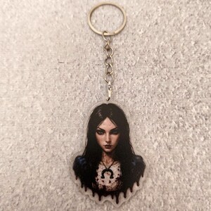 Op de afbeelding: Een doorzichtige acryl sleutelhanger met een illustratie van een persoon met lang donker haar, gekleed in een wit topje met een zwarte hoefijzerhanger. Het ontwerp bevat een zwart druipeffect aan de onderzijde en een zilverkleurige metalen ring.