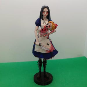 Puede incluir: Figura de un personaje de Alicia en el país de las maravillas, vestida con un vestido azul y un delantal blanco, sosteniendo un cuchillo y una muñeca desmembrada. La figura tiene cabello negro, ojos rojo sangre y salpicaduras de sangre. La base es negra.