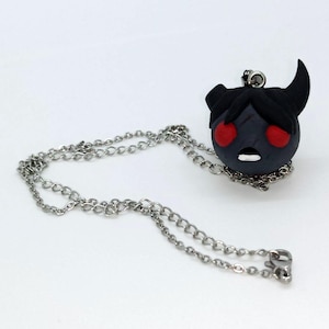 May include: A handmade pendant necklace featuring a dark grey character with red eyes, black hair, and horns. The pendant is attached to a silver-coloured chain. The character has a surprised expression.