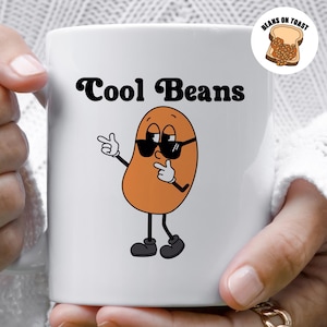 May include: White ceramic mug with a cartoon bean wearing sunglasses and the text "Cool Beans". A small graphic of beans on toast is in the top right corner.