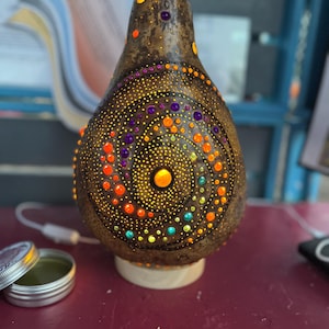 May include: A handcrafted gourd lamp with a brown exterior and a spiral design of colorful dots in orange, yellow, purple, and green. The lamp sits on a small wooden base and is illuminated from within, creating a warm glow.
