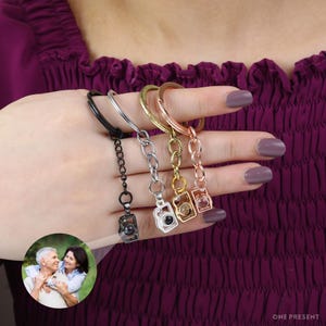 May include: Four silver, gold, rose gold, and black keychains with a camera charm. Each camera charm has a small clear window that reveals a photo inside.