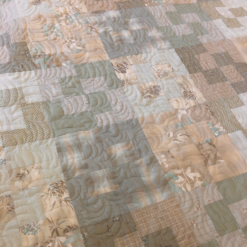 K&ouml;nnte beinhalten: Eine Decke aus verschiedenen Braunt&ouml;nen und Gr&uuml;nt&ouml;nen mit einem gesteppten Muster aus geschwungenen Linien. Die Decke besteht aus Patchwork-Quadraten in verschiedenen Gr&ouml;&szlig;en und Mustern.