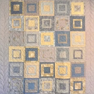 May include: A quilt with a patchwork design featuring squares of different shades of grey, blue, and yellow. The squares are arranged in a grid pattern and are outlined with a darker shade of grey.