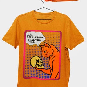 May include: Mustard yellow t-shirt featuring a comic-style illustration of an orange cat holding a skull. The cat is looking at the skull with a speech bubble that reads, "Alas, poor hooman, I knew him well."