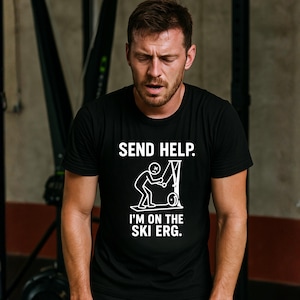 May include: Black t-shirt with white text and graphic. The text reads "SEND HELP." and "I'M ON THE SKI ERG." The graphic depicts a stick figure on a ski erg machine. The person in the image is wearing the shirt.
